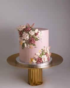 a pink cake decorated with flowers on a gold platter and silver stand against a gray background