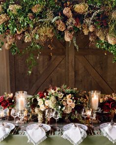 a table topped with lots of flowers and candles next to a wooden wall covered in greenery