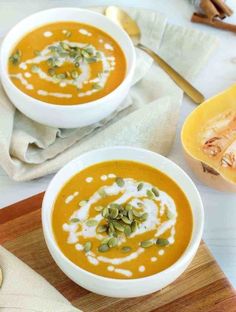 two bowls of pumpkin soup on a wooden cutting board next to a spoon and napkin