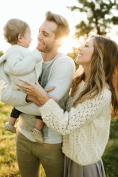 a man and woman holding a baby in their arms while the sun sets behind them