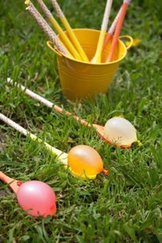 an assortment of colored eggs in the grass next to a yellow bucket with straws