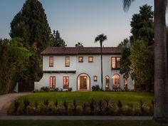 a white house surrounded by palm trees and greenery at dusk with the sun shining through the windows