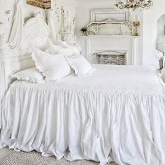 a white bed with ruffled bedspread and pillows on top of it next to a fireplace