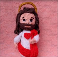 a crocheted jesus doll holding a heart on top of a wooden table next to a pink wall