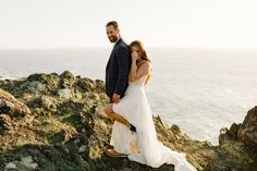 a man and woman standing on top of a rocky cliff next to the ocean with their arms around each other