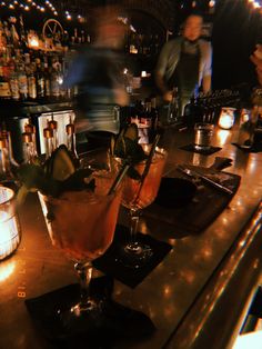 two cocktails sitting on top of a bar next to bottles and glasses filled with liquid