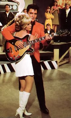 a man holding a guitar while standing next to a woman in a short white dress