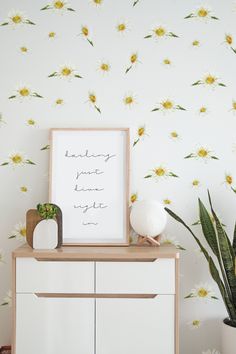 a white dresser topped with a vase filled with flowers next to a wall mounted poster