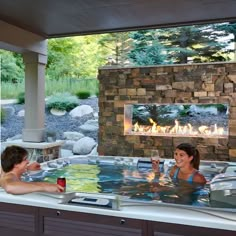 two people sitting in an outdoor hot tub with fire and rocks on the wall behind them