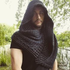 a man wearing a knitted shawl in front of a body of water with trees and grass behind him