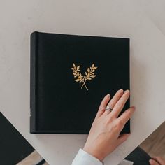 a person's hand on top of a black book with gold leaf decoration, sitting at a table