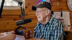 an old man sitting at a table with a cup in front of him and a microphone behind him