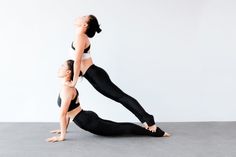 two women doing yoga poses in front of a white wall, one standing and the other sitting on her back