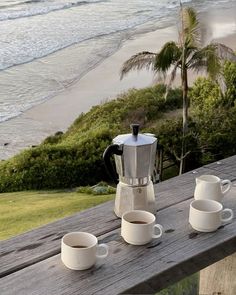 three cups and two coffee makers on a wooden table overlooking the ocean with palm trees in the background