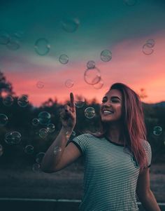 a woman smiles as she holds up soap bubbles in front of an orange and pink sky