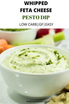 a white bowl filled with pesto dip next to crackers and vegetables on the side