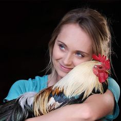 a beautiful young woman holding a rooster in her arms and smiling at the camera while wearing a blue shirt