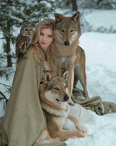 a woman sitting in the snow with two wolfs and an owl on her shoulder