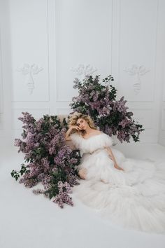 a woman sitting on the floor with flowers in her hair and white dress, surrounded by purple lilacs