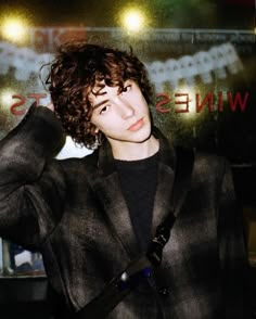 a young man with curly hair wearing a black suit and holding his hand on his head