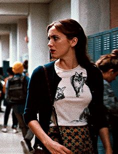 a woman walking down a hallway next to lockers with people in the corridor behind her