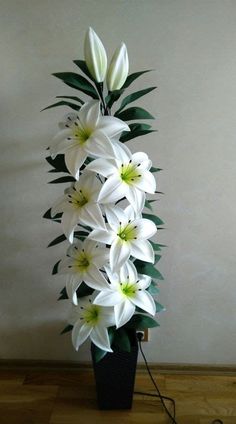 white flowers in a black vase on a wooden floor next to a wall and wires