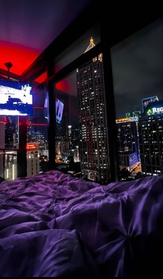 a bed with purple comforter in front of a city skyline at night, lit up by colorful lights