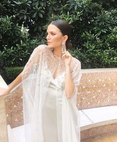 a woman wearing a white dress and veil standing in front of a wall with bushes