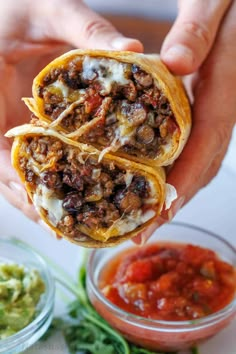 someone is holding two burritos with meat and cheese on them, in front of some guacamole