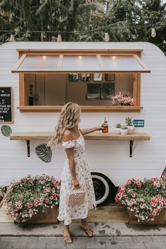 a woman standing in front of a food truck with flowers on the ground and plants behind her