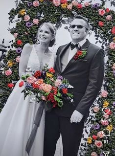 a man and woman standing next to each other in front of a floral arch with roses on it