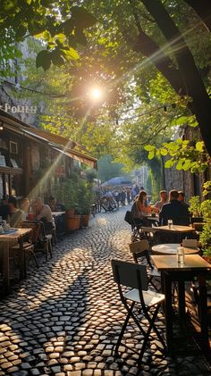 people are sitting at tables in an alleyway with sun shining through the trees and cobblestone pavement