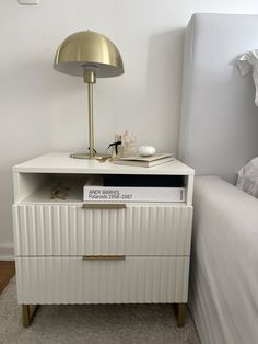 a nightstand with a lamp on top of it next to a white bed in a bedroom