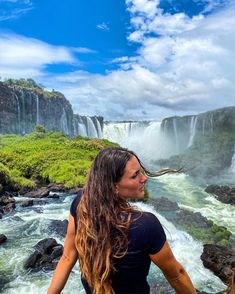 a woman with long hair standing in front of a waterfall and looking at the water