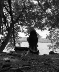 a woman sitting on the ground in front of some trees and looking out at water