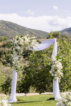 an outdoor wedding ceremony with white flowers on the arch and greenery in the background