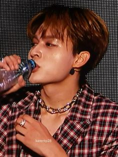 the young man is drinking from a water bottle while wearing a checkered shirt and necklace