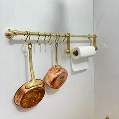 two copper pots are hanging on the wall next to a roll of toilet paper and a pair of brass spoons