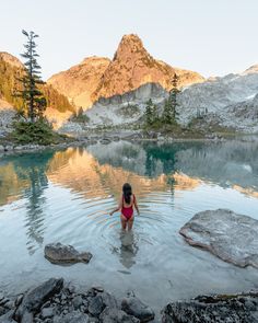 a girl in a red swimsuit wading into a lake with mountains in the background