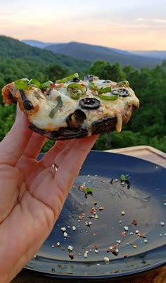 a hand holding up a piece of pizza on top of a blue plate with mountains in the background