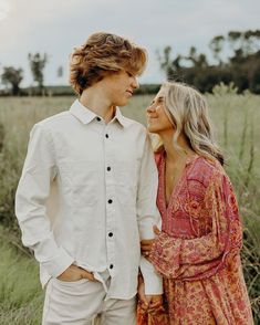 a young man and woman standing next to each other in front of tall grass field