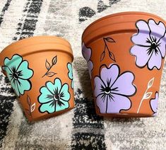 two potted plants sitting on top of a carpet covered in floral designs, one painted orange and the other blue