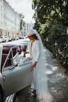 a woman standing next to a white car on the side of a road with flowers in it