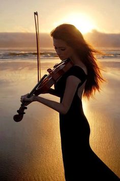 a woman holding a violin on top of a beach next to the ocean at sunset