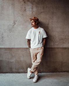 a man leaning against a wall with his hands on his hips wearing a hat and khaki pants