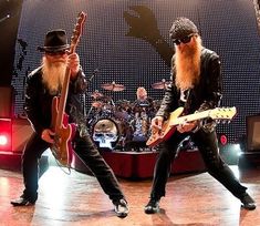 two men with long hair and beards are playing guitars on stage while another man in black wears a hat