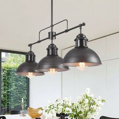 three lights hanging over a dining room table with flowers in the vase on the side