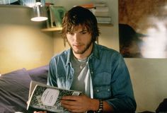 a young man sitting on a bed holding a book in his hands and looking at the camera