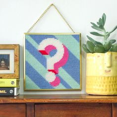 a cross stitch picture hanging on a wall next to a potted plant and two books