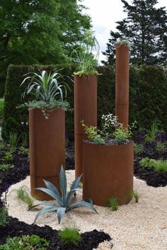 three tall metal planters sitting in the middle of a gravel garden area with plants growing out of them